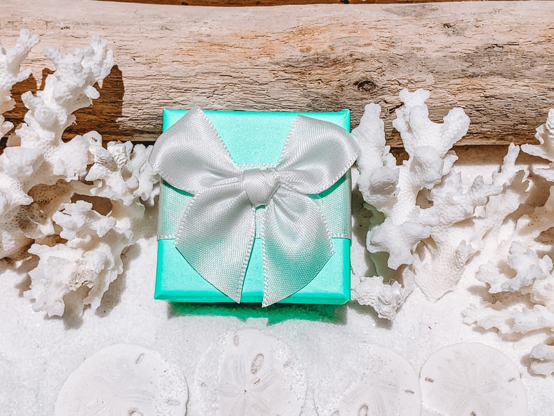 May include: A small, square, turquoise gift box with a white satin bow. The box is surrounded by white coral, sand dollars, and a piece of driftwood, creating a beachy theme. The bow is tied neatly on top of the box.
