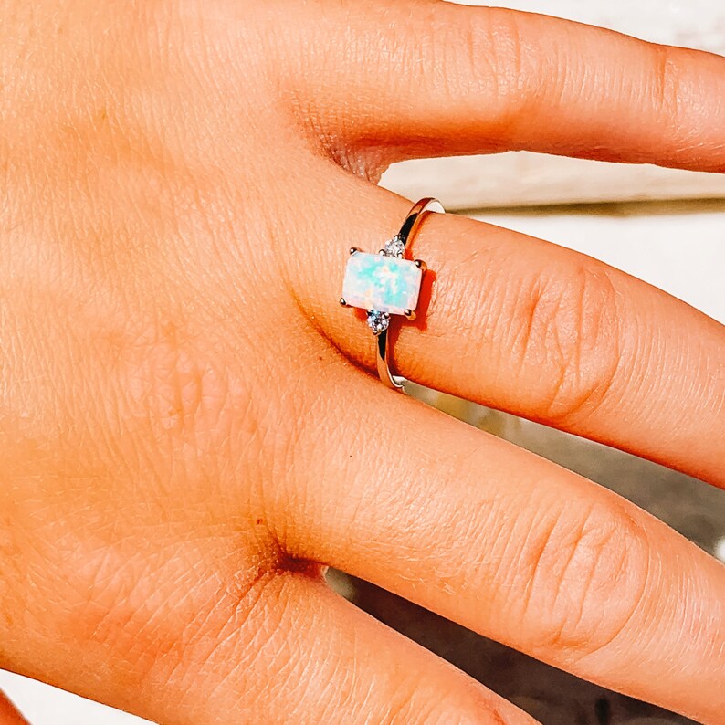 May include: A silver ring with a rectangular opal gemstone. The opal has a light blue and white color. Two small clear gemstones are on either side of the opal. The ring is on a person's finger.