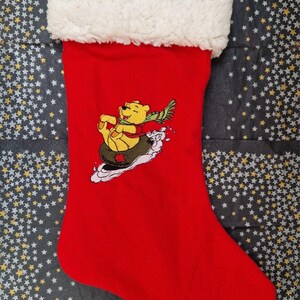 May include: Red Christmas stocking with a white faux fur cuff. The stocking features an embroidered image of Winnie the Pooh ice skating.
