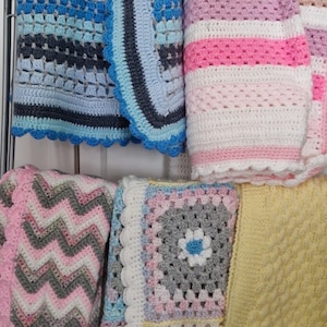 May include: A display of handmade crochet blankets in various colors and patterns. The blankets include blue, pink, white, gray, and yellow. Some have striped designs, while others feature chevron or floral motifs. The blankets are displayed on a metal rack.