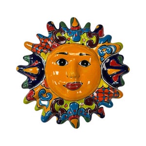 May include: A vibrant, hand-painted ceramic sun face with a smiling expression. The sun features an orange face, red lips, and black eyes. The rays are decorated with colorful patterns in blue, green, yellow, and red. A decorative wall hanging.
