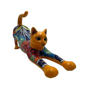 May include: A ceramic cat figurine in a stretching pose. The cat has an orange head and paws, with a body decorated in colorful patterns of blue, red, and green. The tail is curved upwards.