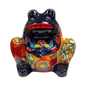 May include: A ceramic frog figurine with a glossy finish. The frog is primarily dark blue with red lips and colorful floral patterns on its body. The figurine has large, expressive eyes and is holding its hands together.