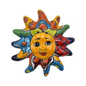 May include: A hand-painted ceramic sun with a smiling face and radiating rays. The sun's face is yellow, with black eyes and a red smiling mouth. The rays are decorated with blue, green, red, and orange patterns.