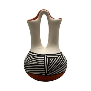 May include: A ceramic wedding vase with a double-spout design connected by a bridge. The base and rims are terracotta-colored, while the upper body is matte white. The rounded lower section features a bold black and white geometric striped pattern.