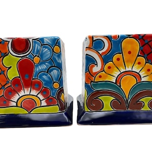 May include: Pair of colorful ceramic bookends. Each bookend features a vibrant floral design with red, orange, yellow, and blue hues. The base is a deep blue, and the bookends are square-shaped.
