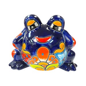 May include: A ceramic frog figurine with a glossy, dark blue finish. The frog features colorful floral patterns in orange, red, yellow, and blue. The frog has large, expressive eyes and a smiling red mouth. A decorative item.