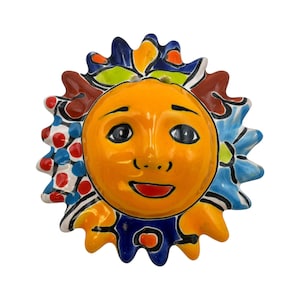 May include: A hand-painted ceramic sun face with a smiling expression. The sun has an orange face, black eyes, and a red mouth. The rays are decorated with blue, red, and white patterns, and the sun's outline is black.