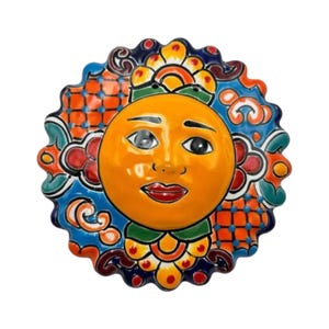 May include: A ceramic sun face wall hanging with a smiling expression. The sun has an orange face, red lips, and detailed eyes. The sun's rays are decorated with blue, orange, and green patterns.