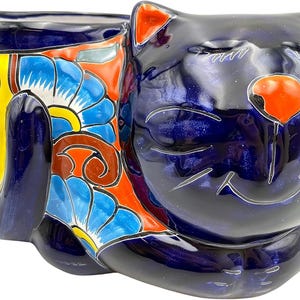May include: A glossy dark blue ceramic cat-shaped planter or container. The cat has a bright orange nose and ear tips, with closed eyes and a smiling mouth. The body features vibrant, multi-colored floral patterns in red, yellow, and blue.
