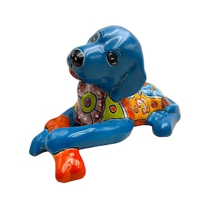 May include: A glossy ceramic dog figurine in a reclining position, featuring a vibrant blue head and limbs. The chest and back are decorated with colorful, intricate patterns in shades of orange, yellow, green, and white, including floral motifs.