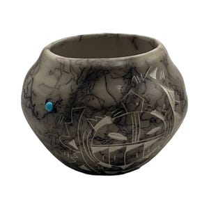 May include: A round, cream-colored ceramic pot featuring a dark, wispy black smoke-fire pattern across its surface. The exterior is decorated with etched geometric line designs and a single, small, round, bright blue turquoise stone inlay.