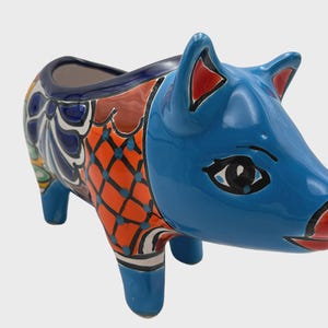 May include: A ceramic pig planter with a vibrant design. The pig is primarily blue with orange and patterned accents. The ears have red details, and the eyes are black. The planter is suitable for small plants or decorative use.
