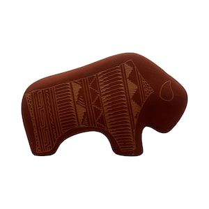 May include: A terracotta-colored bison figurine featuring intricate, etched geometric patterns across its side. The design includes vertical lines, zigzags, and rectangular motifs carved into the surface, with a simple outline of an eye on the head.