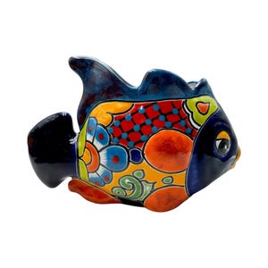 May include: A colorful ceramic fish figurine with a vibrant design. The fish features a blue tail and fins, a dark blue head, and a body adorned with orange, yellow, red, and blue patterns. The fish has a wide opening at the top.