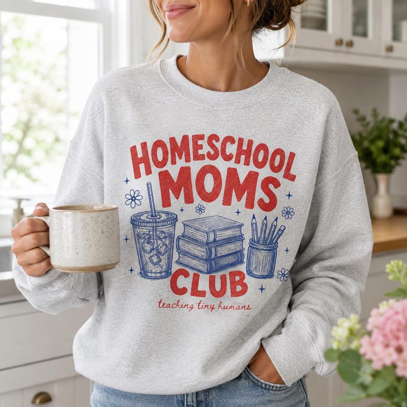 Puede incluir: Una sudadera gris claro de cuello redondo con texto gr&aacute;fico en rojo y azul que dice HOMESCHOOL MOMS CLUB y teaching tiny humans. El dise&ntilde;o incluye un vaso de caf&eacute; helado azul, una pila de tres libros y un portal&aacute;pices con l&aacute;pices.