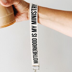 Peut inclure: Lani&egrave;re en tissu blanc avec du texte noir qui dit "MOTHERHOOD IS MY MINISTRY". La lani&egrave;re a un fermoir en m&eacute;tal argent&eacute; et un porte-cl&eacute;s argent&eacute;.