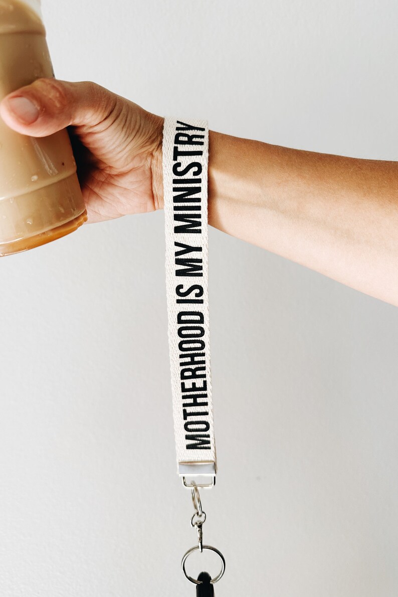 Peut inclure: Lani&egrave;re en tissu blanc avec du texte noir qui dit "MOTHERHOOD IS MY MINISTRY". La lani&egrave;re a un fermoir en m&eacute;tal argent&eacute; et un porte-cl&eacute;s argent&eacute;.