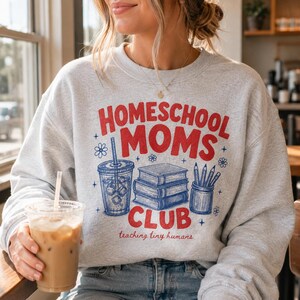Puede incluir: Una sudadera gris claro de cuello redondo con el texto rojo "HOMESCHOOL MOMS CLUB" y el texto azul "teaching tiny humans". El gr&aacute;fico incluye una ilustraci&oacute;n de l&iacute;nea azul de una bebida con pajita, una pila de tres libros y un portal&aacute;pices.