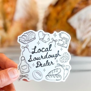 K&ouml;nnte beinhalten: Ein wei&szlig;er Aufkleber mit schwarzen Strichzeichnungen von Backutensilien und Brot. Der Text "Local Sourdough Dealer" ist in einer handschriftlichen Schrift geschrieben. Der Aufkleber wird von einer Hand gehalten, mit einem unscharfen Hintergrund.