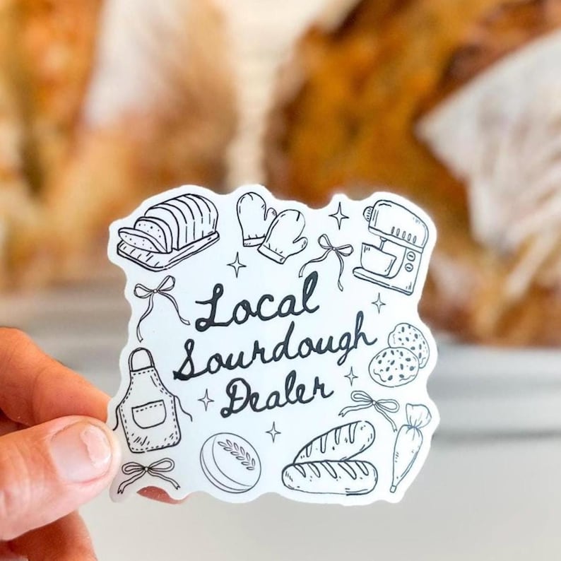 K&ouml;nnte beinhalten: Ein wei&szlig;er Aufkleber mit schwarzen Strichzeichnungen von Backutensilien und Brot. Der Text "Local Sourdough Dealer" ist in einer handschriftlichen Schrift geschrieben. Der Aufkleber wird von einer Hand gehalten, mit einem unscharfen Hintergrund.