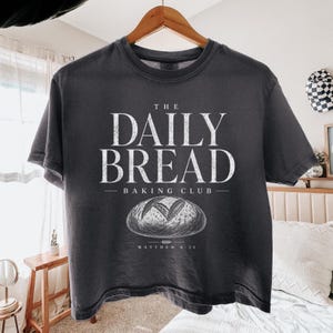 Może przedstawiać: Ciemnoszary t-shirt z krótkim rękawem z białym, postarzanym napisem THE DAILY BREAD BAKING CLUB, grafiką przedstawiającą bochenek chleba oraz tekstem MATTHEW 6:11 nadrukowanym pod ilustracją chleba na przedniej części koszulki.