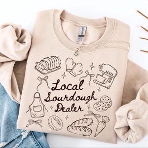 May include: A beige sweatshirt with a black graphic design featuring bread, baking tools, and the text "Local Sourdough Dealer." A silver necklace with a sun pendant is also visible. The sweatshirt is folded on top of blue denim.