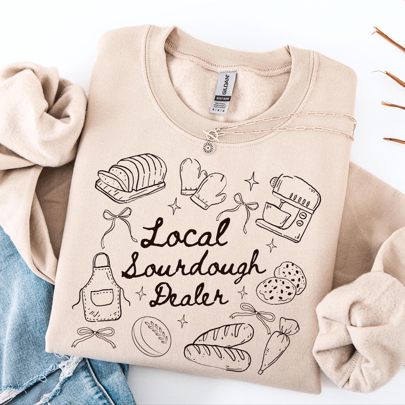 May include: A beige sweatshirt with a black graphic design featuring bread, baking tools, and the text "Local Sourdough Dealer." A silver necklace with a sun pendant is also visible. The sweatshirt is folded on top of blue denim.