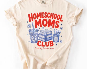 Puede incluir: Una camiseta de color crema con texto rojo que dice HOMESCHOOL MOMS CLUB y texto rojo más pequeño que dice teaching tiny humans. El gráfico incluye dibujos de línea azul de una bebida con pajita, tres libros apilados y un portalápices.