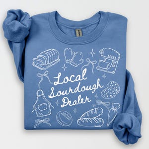 May include: A light blue sweatshirt with white line art of baking tools and bread. The text "Local Sourdough Dealer" is written in white script. The design includes a mixer, bread, and oven mitts.