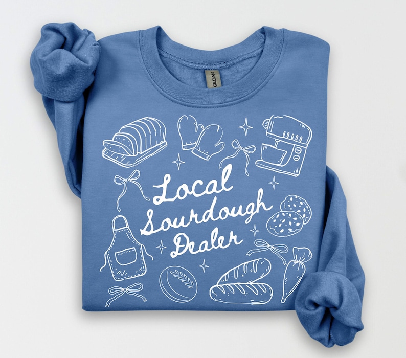 May include: A light blue sweatshirt with white line art of baking tools and bread. The text "Local Sourdough Dealer" is written in white script. The design includes a mixer, bread, and oven mitts.