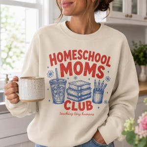 Puede incluir: Una sudadera de cuello redondo color crema con un estampado gr&aacute;fico en rojo y azul. El texto dice "HOMESCHOOL MOMS CLUB" y "teaching tiny humans" con ilustraciones de una bebida, una pila de tres libros y un portal&aacute;pices.