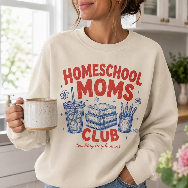 Puede incluir: Una sudadera de cuello redondo color crema con un estampado gr&aacute;fico en rojo y azul. El texto dice "HOMESCHOOL MOMS CLUB" y "teaching tiny humans" con ilustraciones de una bebida, una pila de tres libros y un portal&aacute;pices.