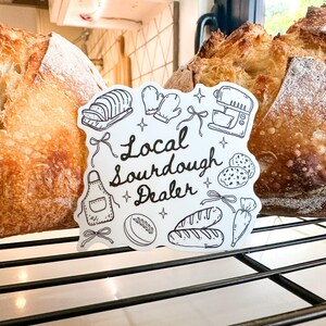 K&ouml;nnte beinhalten: Zwei Laibe frisch gebackenes Sauerteigbrot mit einem Schild mit der Aufschrift "Local Sourdough Dealer". Das Schild zeigt Illustrationen von Backwerkzeugen und Zutaten. Das Brot befindet sich auf einem Metallrost.