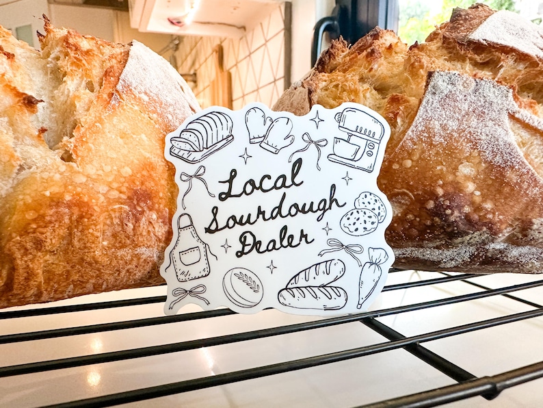 K&ouml;nnte beinhalten: Zwei Laibe frisch gebackenes Sauerteigbrot mit einem Schild mit der Aufschrift "Local Sourdough Dealer". Das Schild zeigt Illustrationen von Backwerkzeugen und Zutaten. Das Brot befindet sich auf einem Metallrost.