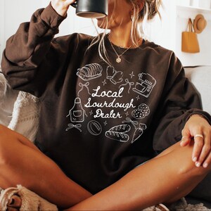 May include: A dark brown sweatshirt with the text "Local Sourdough Dealer" and white illustrations of baking tools and bread. The design includes an apron, oven mitts, and a mixer.