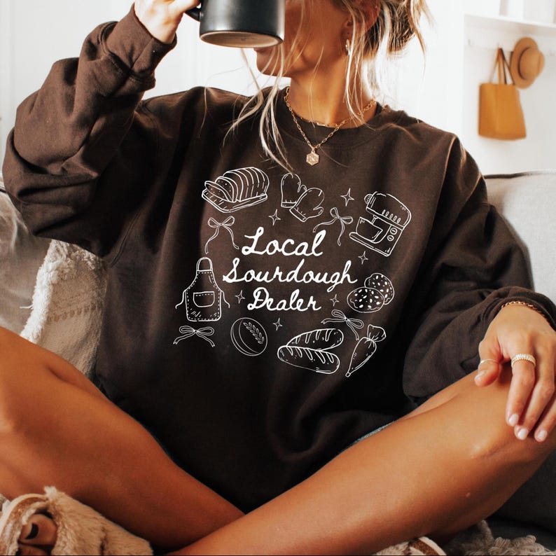 May include: A dark brown sweatshirt with the text "Local Sourdough Dealer" and white illustrations of baking tools and bread. The design includes an apron, oven mitts, and a mixer.