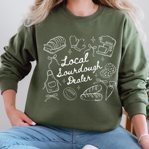 May include: An olive green sweatshirt with a white graphic design featuring baking-related illustrations, including bread, oven mitts, and a mixer. The text "Local Sourdough Dealer" is also included in the design.