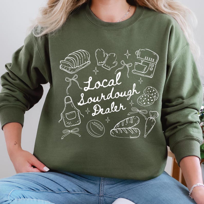 May include: An olive green sweatshirt with a white graphic design featuring baking-related illustrations, including bread, oven mitts, and a mixer. The text "Local Sourdough Dealer" is also included in the design.