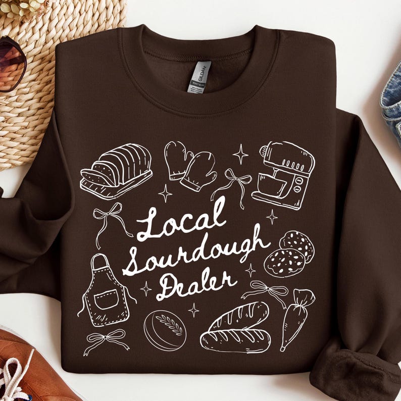 May include: A brown sweatshirt with a white graphic design featuring bread, oven mitts, a mixer, and the text "Local Sourdough Dealer". The design includes stars and bows, creating a bakery theme.