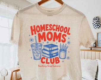 May include: A cream-coloured t-shirt featuring a graphic with the red text "HOMESCHOOL MOMS CLUB" and the blue text "teaching tiny humans". The design includes a blue iced coffee cup, a stack of three books, and a pencil holder with pencils.
