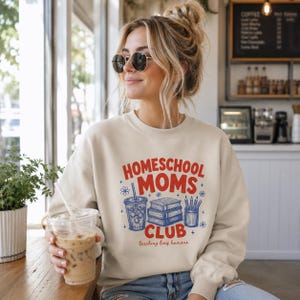 Puede incluir: Una sudadera beige de cuello redondo con un estampado gr&aacute;fico que incluye el texto rojo "HOMESCHOOL MOMS CLUB" y el texto rojo m&aacute;s peque&ntilde;o "teaching tiny humans". El dise&ntilde;o incluye ilustraciones azules de un vaso, libros y l&aacute;pices.