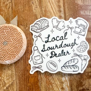 K&ouml;nnte beinhalten: Ein Brotmesser aus Holz mit einem dekorativen Muster und einem Aufkleber mit der Aufschrift "Local Sourdough Dealer" mit Illustrationen von Brot, Backutensilien und Zutaten. Die Artikel befinden sich auf einer Holzoberfl&auml;che.