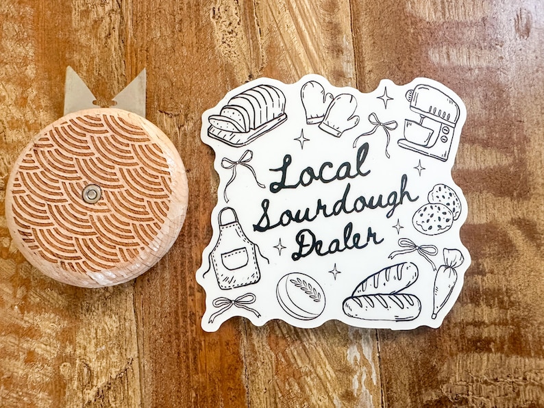 K&ouml;nnte beinhalten: Ein Brotmesser aus Holz mit einem dekorativen Muster und einem Aufkleber mit der Aufschrift "Local Sourdough Dealer" mit Illustrationen von Brot, Backutensilien und Zutaten. Die Artikel befinden sich auf einer Holzoberfl&auml;che.