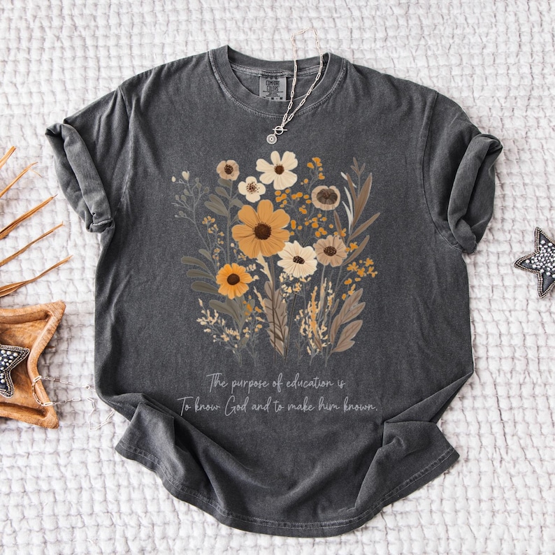 May include: A dark gray t-shirt with a floral design in shades of brown, yellow, and white. The text "The purpose of education is to know God and to make him known." is printed on the bottom of the shirt.