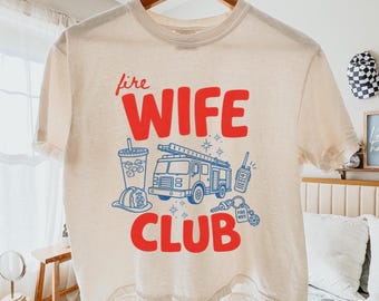 Puede incluir: Una camiseta de manga corta color crema con texto rojo que dice "fire WIFE CLUB" y gráficos de línea azul de un camión de bomberos, un vaso, un casco de bombero, un walkie-talkie y un llavero con una etiqueta que dice "FIRE WIFE".