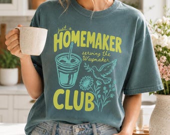 May include: A slate blue short-sleeve t-shirt featuring a graphic print in lime green and light blue. The text reads "just a HOMEMAKER serving the Waymaker CLUB" alongside illustrations of a drink in a cup, a dove, and wildflowers.