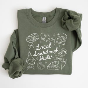 May include: Olive green sweatshirt with white graphic illustrations of bread, oven mitts, a mixer, and other baking-related items. The text "Local Sourdough Dealer" is written in a script font.