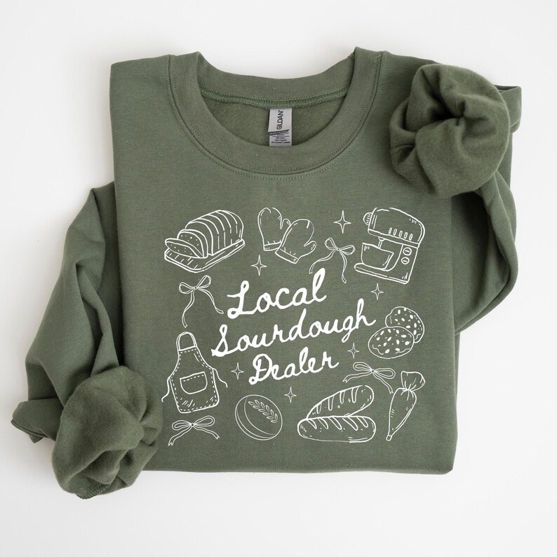 May include: Olive green sweatshirt with white graphic illustrations of bread, oven mitts, a mixer, and other baking-related items. The text "Local Sourdough Dealer" is written in a script font.