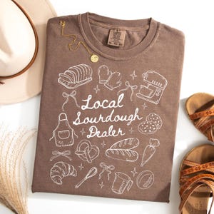 May include: A brown Comfort Colors t-shirt with a white graphic design featuring baking-related illustrations and the text "Local Sourdough Dealer." The shirt is adorned with a gold necklace and is paired with sandals and a hat.